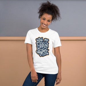 May include: A white t-shirt with the text "Ask me about my current hyper fixation" in a navy blue and orange design. The shirt is made of a soft material and has short sleeves.
