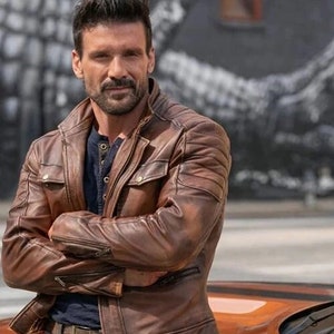 May include: A man wearing a brown leather jacket with a zipper closure and a button-up collar. He is standing with his arms crossed in front of a yellow car.