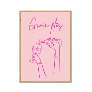 Könnte beinhalten: Gerahmter rosa Kunstdruck mit einer Strichzeichnung von Händen, die Wein in ein Glas gießen. Die Worte "Gwin plis" sind in kursiver Schrift geschrieben. Das Kunstwerk hat einen minimalistischen Stil. Der Rahmen ist aus Holz.