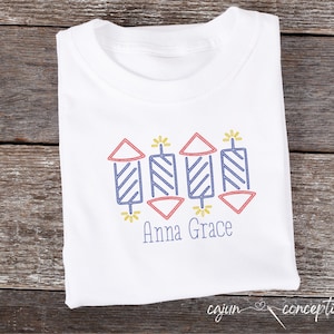 May include: White t-shirt with a red, blue, and yellow embroidered design of fireworks and the name "Anna Grace" underneath.