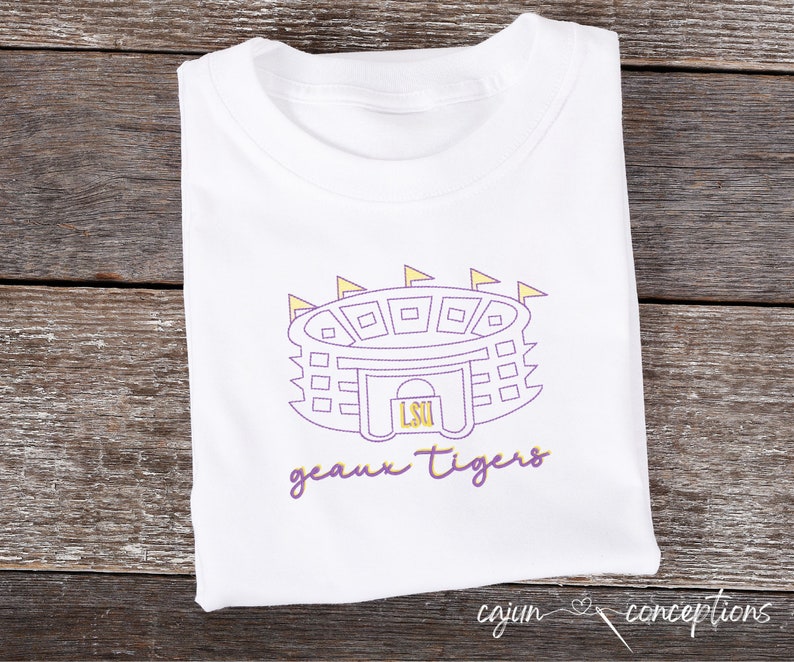 K&ouml;nnte beinhalten: Wei&szlig;es T-Shirt mit einer lila und gelben Umrandung eines Stadions mit dem Text "LSU" im Inneren. Der Text "geaux tigers" befindet sich unter dem Stadion.