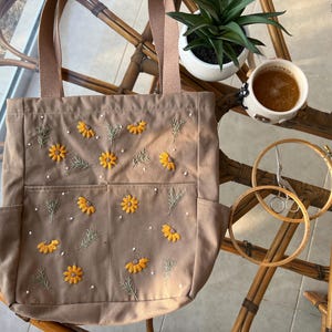 May include: A brown canvas tote bag with embroidered yellow flowers and white beads. The bag has three pockets on the front.