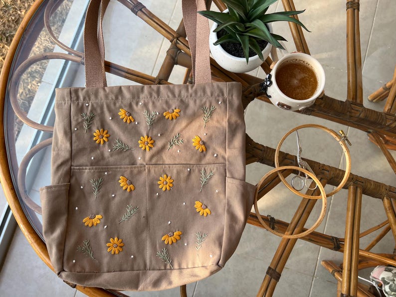 May include: A brown canvas tote bag with embroidered yellow flowers and white beads. The bag has three pockets on the front.