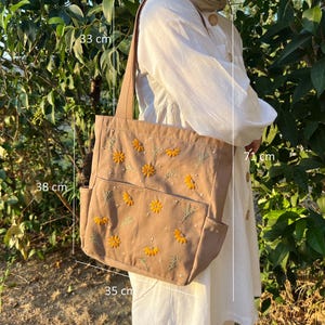 May include: A brown canvas tote bag with embroidered yellow flowers and green leaves. The bag has a top zipper closure and two side pockets. The bag measures 38 cm wide, 33 cm tall, and 35 cm deep.