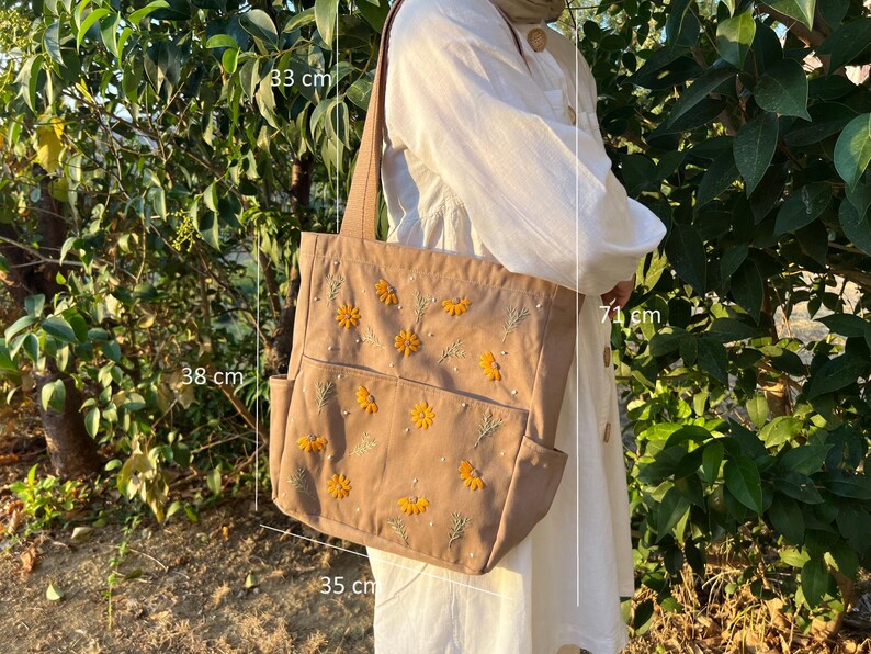 May include: A brown canvas tote bag with embroidered yellow flowers and green leaves. The bag has a top zipper closure and two side pockets. The bag measures 38 cm wide, 33 cm tall, and 35 cm deep.