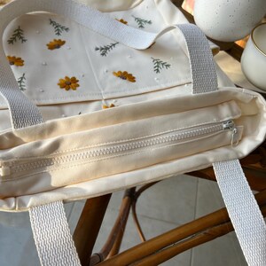 May include: A cream-coloured canvas tote bag with embroidered flowers and leaves. The bag has a zipper closure and two white straps.