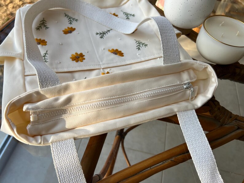 May include: A cream-coloured canvas tote bag with embroidered flowers and leaves. The bag has a zipper closure and two white straps.