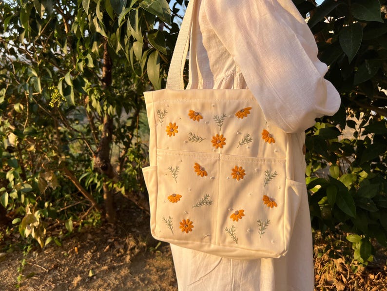 May include: A white canvas tote bag with embroidered yellow flowers and green leaves. The bag has multiple pockets.