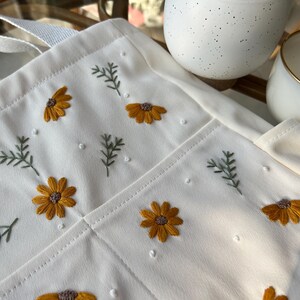 May include: A white canvas tote bag with embroidered yellow flowers and green leaves. The bag has a single strap and a pocket on the front.