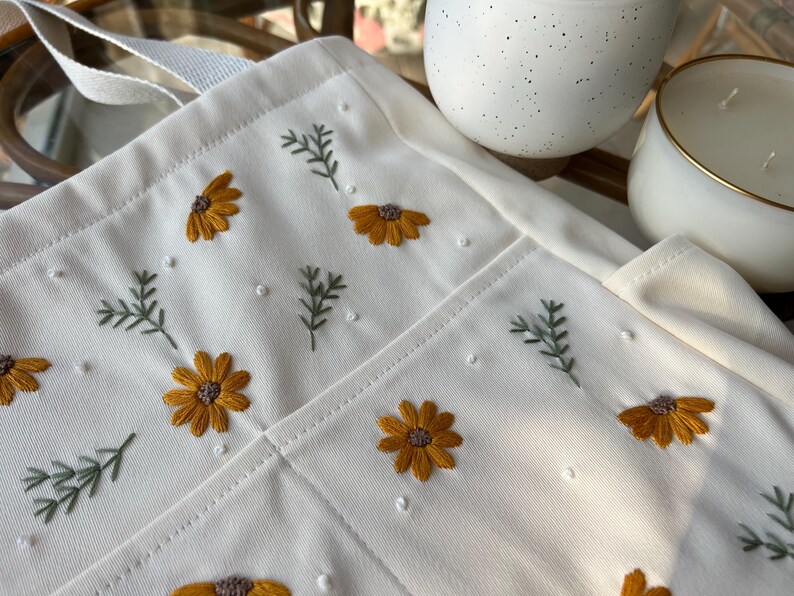 May include: A white canvas tote bag with embroidered yellow flowers and green leaves. The bag has a single strap and a pocket on the front.