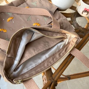 May include: A brown canvas tote bag with a zipper closure and a light brown lining. The bag has embroidered flowers and leaves in yellow and green.