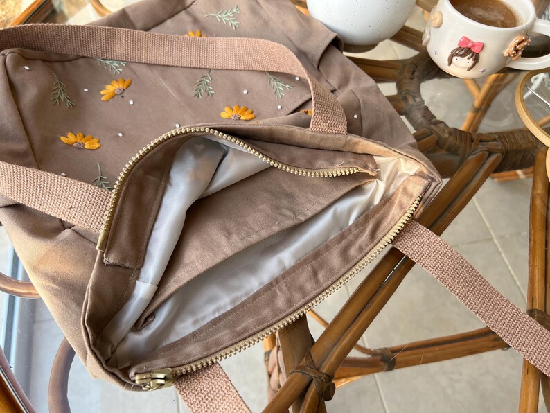 May include: A brown canvas tote bag with a zipper closure and a light brown lining. The bag has embroidered flowers and leaves in yellow and green.