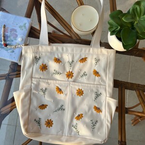 May include: A white canvas tote bag with embroidered yellow flowers and white pearls. The bag has two pockets on the front and two handles.