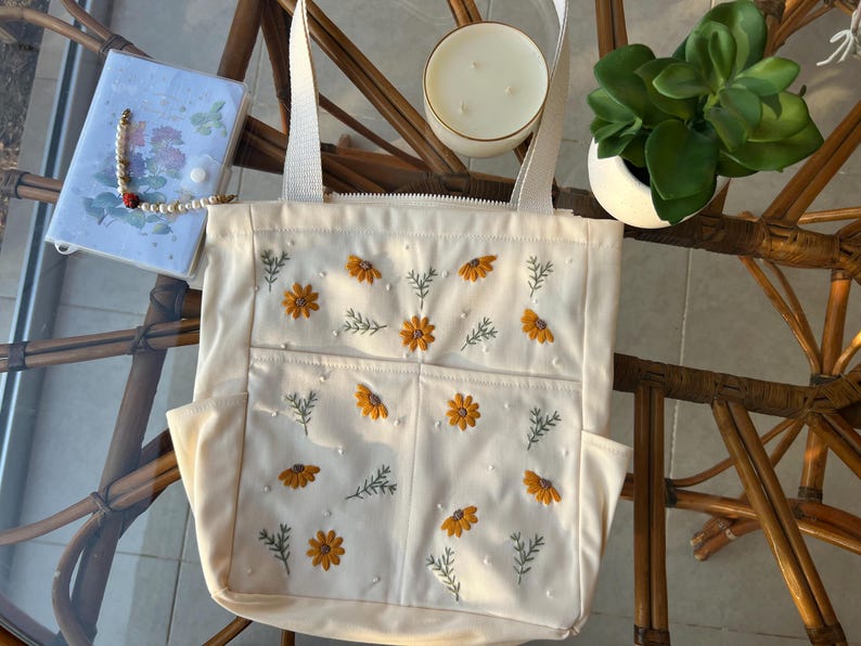 May include: A white canvas tote bag with embroidered yellow flowers and white pearls. The bag has two pockets on the front and two handles.