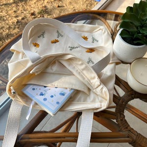 May include: A white canvas tote bag with embroidered flowers and a zip closure. The bag is open and a small notebook with a blue and white pattern is inside.
