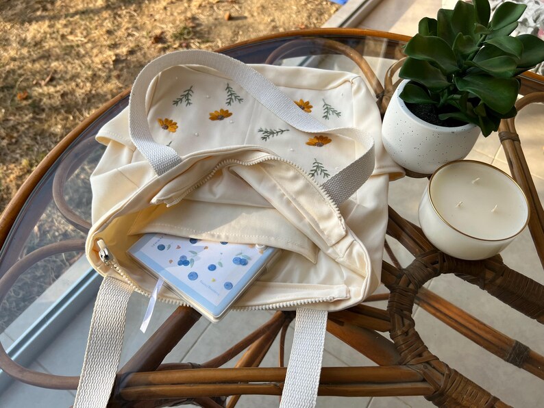 May include: A white canvas tote bag with embroidered flowers and a zip closure. The bag is open and a small notebook with a blue and white pattern is inside.
