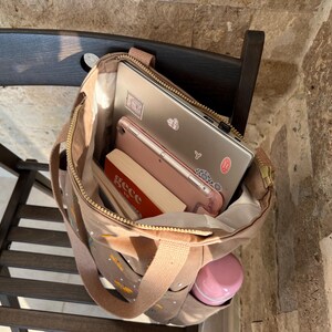 May include: A brown canvas tote bag with gold zipper accents and a gold zipper pull. The bag is filled with a laptop, a tablet, a book, and a pink water bottle.