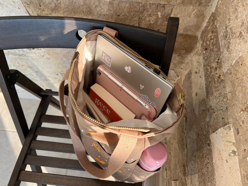 May include: A brown canvas tote bag with gold zipper accents and a gold zipper pull. The bag is filled with a laptop, a tablet, a book, and a pink water bottle.