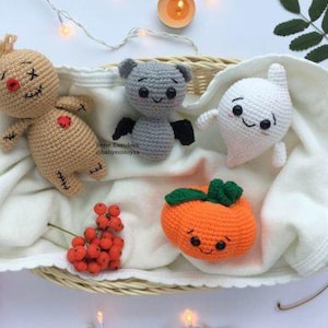May include: A selection of handmade crochet Halloween toys, including a scarecrow, a bat, a ghost, and a pumpkin, presented in a woven basket on a white cloth. The toys are in beige, grey, white, and orange, with black embroidered details.