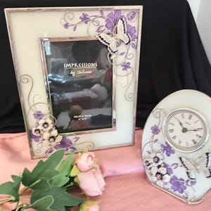 May include: A decorative set featuring a photo frame and a clock. The frame and clock have a white background with silver trim, adorned with purple floral and butterfly accents. The frame has the text "IMPRESSIONS by Juliane".