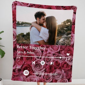 Pode incluir: Um cobertor rosa e branco com um padrão de rosas. O cobertor apresenta uma foto de um casal se beijando, uma interface de reprodutor de música e o texto "Better Together Alex & John".