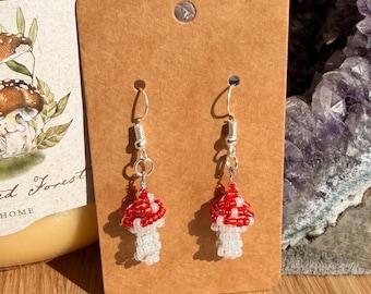 May include: A pair of dangle earrings featuring mushroom-shaped charms crafted from small red and white glass beads. The earrings hang from silver-toned metal hooks and are displayed on a rectangular brown cardboard backing card with a hole at the top.