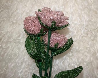 May include: A decorative floral arrangement featuring three roses crafted from small, light pink glass beads. Each rose is attached to a green stem wrapped in tape, accompanied by several detailed green leaves also constructed from tiny glass beads.