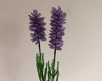 May include: Two decorative floral stems crafted from small purple and green glass beads. Each stem features a long, textured purple beaded flower head atop a thin, dark green wire stalk adorned with multiple slender, green beaded leaves.