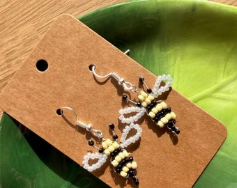 May include: A pair of dangle earrings featuring bee-shaped designs crafted from small yellow, black, and translucent white seed beads. The bees have striped bodies and looped wings, attached to silver-toned metal fishhook ear wires on a brown card.