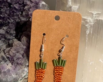 May include: A pair of dangle earrings shaped like carrots, crafted from small orange and green glass beads. The earrings feature silver-toned metal hooks and are displayed on a rectangular brown cardboard backing card with a hole at the top.