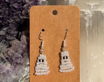 May include: A pair of dangle earrings featuring ghost-shaped charms crafted from small white seed beads with two black seed beads for eyes. The earrings are attached to a rectangular brown cardboard display card with a hole at the top for hanging.