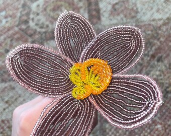May include: A handcrafted flower sculpture made from small, metallic mauve-colored beads forming five distinct petals. The center of the flower features a cluster of bright yellow and orange beads, creating a vibrant contrast against the mauve petals.