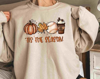 May include: A beige sweatshirt with a graphic design of a pumpkin, a baseball, a leaf, and a coffee cup. The text "'Tis the season" is printed below the graphic.