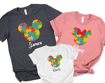 May include: Three t-shirts with a puzzle piece Mickey Mouse design. The shirts are in navy blue, pink, and white. The shirts have the names "James", "Mary", and "David" printed on them.