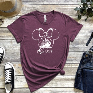 Op de afbeelding: Bordeauxkleurig T-shirt met een witte omtrek van Mickey Mouse oren met een strik, een cruiseschip, een anker en de tekst "2024".