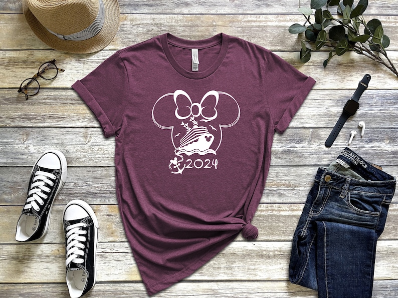 Op de afbeelding: Bordeauxkleurig T-shirt met een witte omtrek van Mickey Mouse oren met een strik, een cruiseschip, een anker en de tekst "2024".