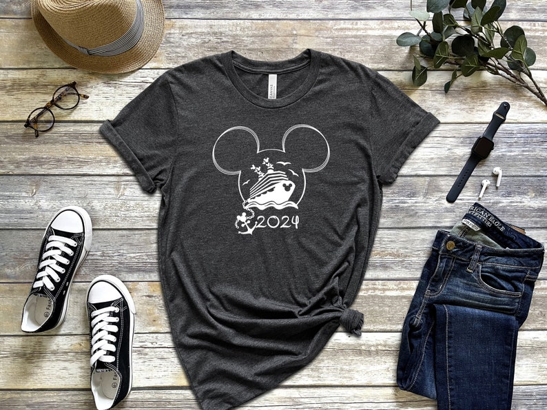Op de afbeelding: Een donkergrijs T-shirt met een wit Mickey Mouse silhouetontwerp. Binnen de oren bevindt zich een cruiseschip met palmbomen en de tekst "2024".