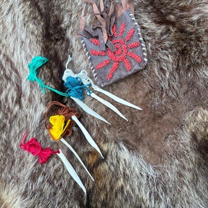 May include: A small, gray pouch with a red sunburst design made of beads, and white stitching. Several bundles of colorful yarn and white, pointed tools are arranged around the pouch on a brown, furry surface.