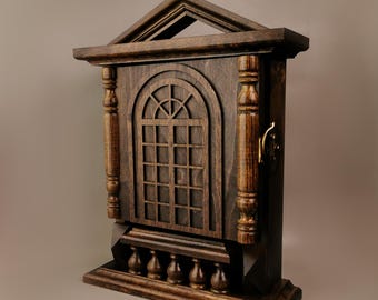 Op de afbeelding: Een donkerbruine houten sleutelhanger in de vorm van een huis met een decoratief raam en een klein hek. De sleutelhanger heeft een messing sluiting.