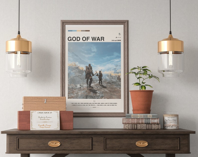 K&ouml;nnte beinhalten: Ein gerahmtes Poster mit dem Titel "God of War" und dem Erscheinungsdatum "20. April 2018". Das Poster zeigt eine Videospielszene mit zwei Charakteren, die durch eine schneebedeckte Landschaft gehen. Das Poster h&auml;ngt an einer Wand &uuml;ber einem Holztisch mit einer Topfpflanze, B&uuml;chern und einer Kerze.