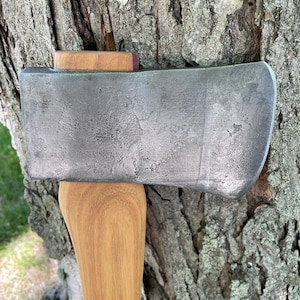 Puede incluir: Primer plano de una cabeza de hacha gris con mango de madera. El hacha está apoyada en un tronco de árbol.