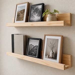 Könnte beinhalten: Zwei horizontale, hellfarbene Holzregale, die an einer Wand montiert sind. Das obere Regal enthält zwei gerahmte Bilder und eine kleine Topfpflanze. Das untere Regal zeigt drei Bücher und zwei gerahmte Bilder, die linear angeordnet sind.