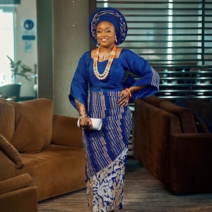 May include: A woman wearing a blue and white patterned dress with a blue head wrap and a beaded necklace. She is holding a white clutch purse and standing in a room with brown couches.