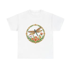 May include: White t-shirt featuring a detailed illustration of a dragonfly within a circular floral design. The dragonfly has brown wings and a green body, surrounded by orange and purple flowers and green leaves. The design is framed by a gold border.