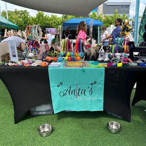 May include: A black table covered with a turquoise tablecloth with the text "Anita's Stitches" in white. The table is set up at an outdoor market and is filled with colorful dog collars, leashes, and toys. There are two silver dog bowls on the green grass in front of the table.