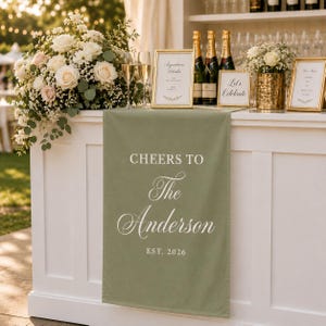 Könnte beinhalten: Ein salbeigrüner Tischläufer aus Stoff mit weißem Aufdruck CHEERS TO The Anderson EST. 2026. Der Läufer hängt über der Vorderseite einer weißen Bartheke, unterhalb eines Blumenarrangements, Champagnerflaschen und gerahmten Schildern.