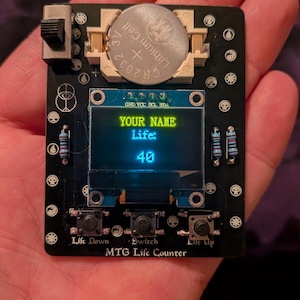 May include: Black MTG Life Counter with a screen displaying "YOUR NAME" and "Life: 40". It has three buttons labeled "Lift Down", "Switch", and "Life Up". A CR2032 3V battery is visible.