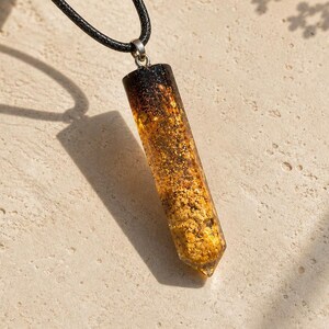 Puede incluir: Un collar con colgante que presenta un colgante rectangular de resina color ámbar con motas doradas, suspendido de un cordón negro. El colgante tiene una parte superior marrón oscuro y una base puntiaguda. El collar se muestra sobre una superficie beige claro.