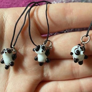 Puede incluir: Tres pequeños colgantes de oso panda. Cada panda es blanco con detalles negros en las orejas, los ojos y los pies. Los colgantes están ensartados en cordones negros con bucles de metal plateado.