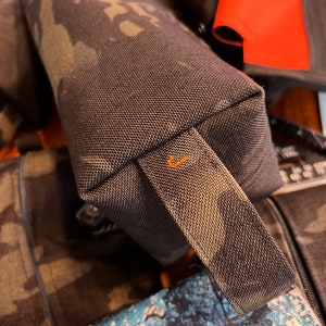 Pode incluir: Um estojo para lápis de tecido camuflado preto e verde com um zíper preto e uma alça de tecido camuflado preto e verde. A alça tem um detalhe de costura laranja.